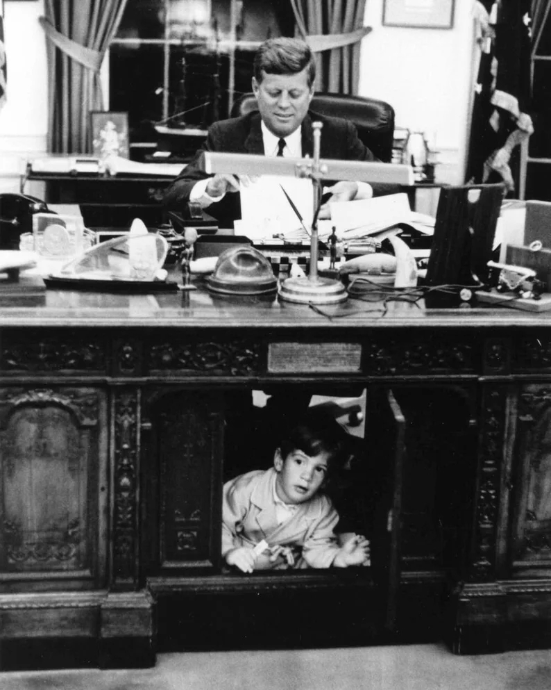 Image President John F Kennedy working at his desk in the Oval Office with his son. John, Jr.